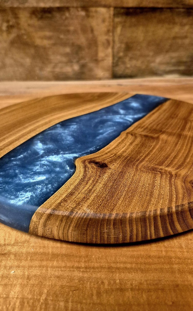 May include: A round wooden serving board with a dark blue epoxy resin river design. The wood has a warm, natural tone, and the resin has a glossy finish. The board is ideal for serving cheese, charcuterie, or other appetizers.