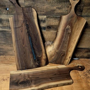 May include: Three handcrafted wooden cutting boards with natural edges. The boards are made of dark brown wood with unique grain patterns. Each board has a handle with a circular opening, perfect for kitchen use.