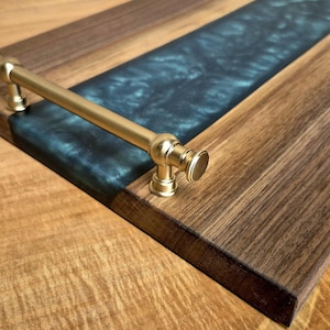 Puede incluir: Una bandeja rectangular de madera con un centro de resina azul oscuro y asas de latón. La bandeja está hecha de madera oscura con un patrón de vetas natural, y las asas están unidas a los lados.