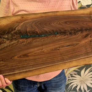 May include: A rectangular wooden serving board with a live edge. The board is a rich brown color with a teal-colored resin inlay. The wood grain is visible throughout the board. The board is being held by a person.