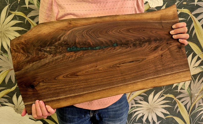 May include: A rectangular wooden serving board with a live edge. The board is a rich brown color with a teal-colored resin inlay. The wood grain is visible throughout the board. The board is being held by a person.