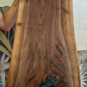 May include: A handcrafted wooden serving board with a live edge design. The board features a rich, dark brown wood grain with a teal-colored resin inlay. The board is held up by a person.