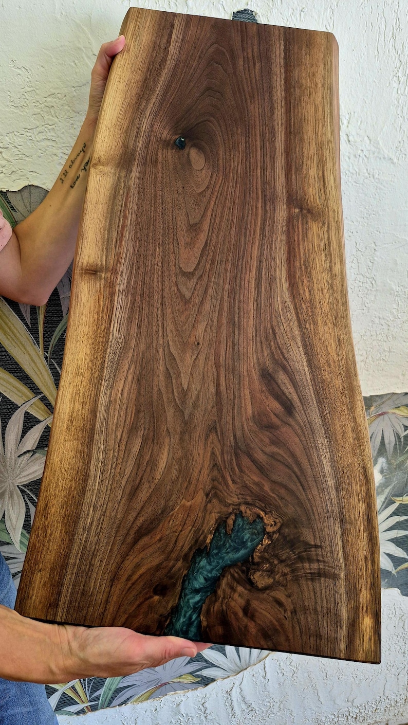 May include: A handcrafted wooden serving board with a live edge design. The board features a rich, dark brown wood grain with a teal-colored resin inlay. The board is held up by a person.