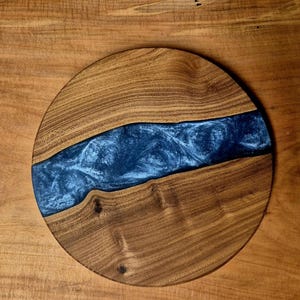 May include: A round wooden serving board with a river of blue resin. The wood has a natural grain and a warm brown color. The resin has a swirling, textured appearance, creating a unique decorative piece.
