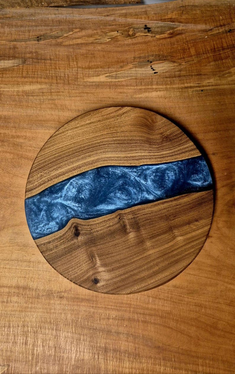 May include: A round wooden serving board with a river of blue resin. The wood has a natural grain and a warm brown color. The resin has a swirling, textured appearance, creating a unique decorative piece.