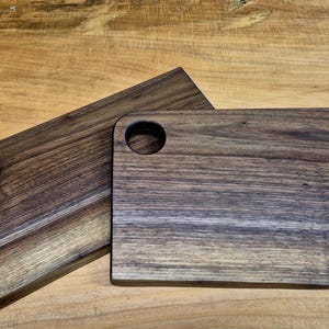 May include: Two rectangular wooden cutting boards with rounded corners and a circular handle hole. The boards have a dark brown color with visible wood grain. They are displayed on a light brown wooden surface.