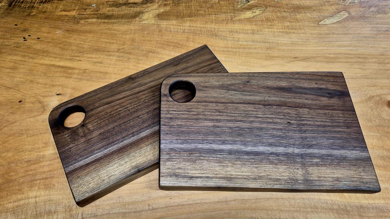 May include: Two rectangular wooden cutting boards with rounded corners and a circular handle hole. The boards have a dark brown color with visible wood grain. They are displayed on a light brown wooden surface.