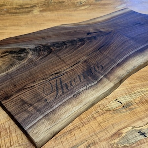 May include: A handcrafted wooden serving board with a natural edge and a handle. The dark brown wood is engraved with the name "Thomas" and "est. 2017". The board is displayed on a light brown wooden surface.