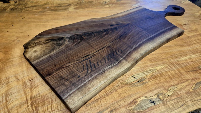May include: A handcrafted wooden serving board with a natural edge and a handle. The dark brown wood is engraved with the name "Thomas" and "est. 2017". The board is displayed on a light brown wooden surface.
