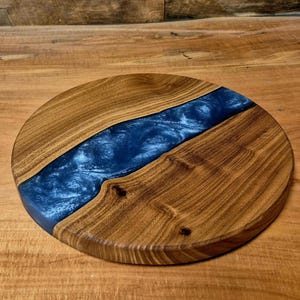 May include: Round wooden serving board with a river of blue resin. The board is made of light brown wood with a dark blue resin center. The wood grain is visible, and the board has a smooth finish.