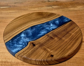 May include: Circular wooden serving board featuring a blue resin river. The board is crafted from light brown wood, with a dark blue resin inlay. The wood grain is visible, and the board has a smooth finish.