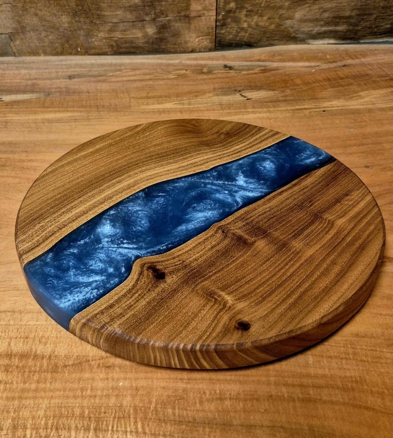 May include: Round wooden serving board with a river of blue resin. The board is made of light brown wood with a dark blue resin center. The wood grain is visible, and the board has a smooth finish.