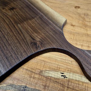 May include: A wooden cutting board with a handle. The board is made of dark brown wood with a natural grain pattern. The handle has a circular opening. The board is on a light brown wooden surface.