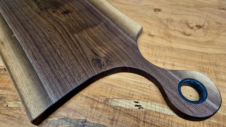 May include: A wooden cutting board with a handle. The board is made of dark brown wood with a natural grain pattern. The handle has a circular opening. The board is on a light brown wooden surface.