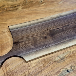 May include: A rectangular wooden cutting board with a handle. The board is made of dark brown wood with a natural edge and a smooth, polished surface. The handle has a circular opening.