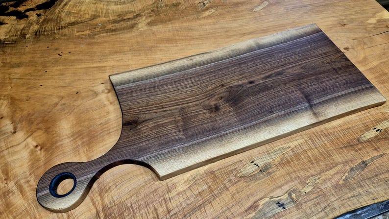May include: A rectangular wooden cutting board with a handle. The board is made of dark brown wood with a natural edge and a smooth, polished surface. The handle has a circular opening.