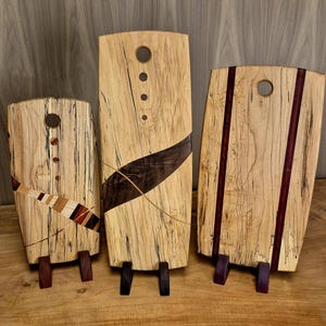 May include: Three wooden cutting boards with decorative designs. The boards are light-colored with dark wood accents and small feet. One board has a diagonal inlay, another has vertical stripes, and the third has a curved inlay.