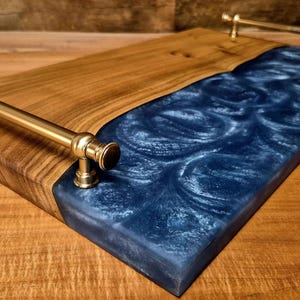 Puede incluir: Una bandeja de madera rectangular con una incrustación de resina azul oscuro. La bandeja tiene asas de latón y un acabado de vetas de madera natural en un lado. La resina tiene un aspecto arremolinado y texturizado.