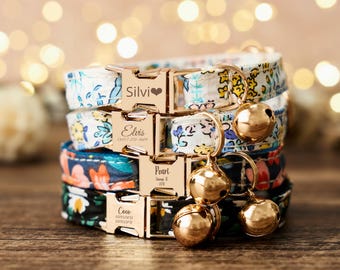 Puede incluir: Una pila de cuatro collares para gatos con estampados florales en azul, blanco y negro. Cada collar tiene una hebilla dorada grabada con un nombre y una campana. Los nombres son Silvi, Elvis, Pearl y Coco.