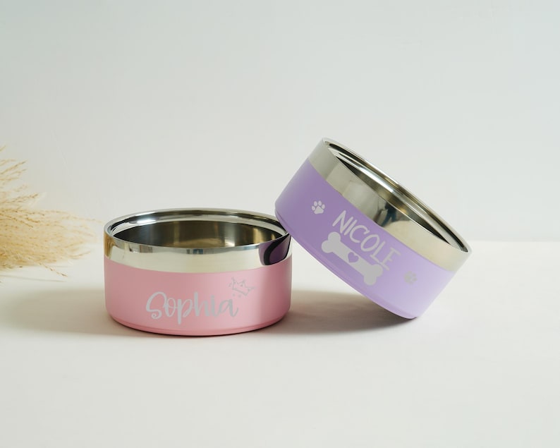 May include: Two stainless steel pet bowls, one pink and one lavender, with silver rims. The pink bowl has the name "Sophia" and a crown graphic. The lavender bowl has the name "Nicole" and a bone graphic. Both bowls are for pets.