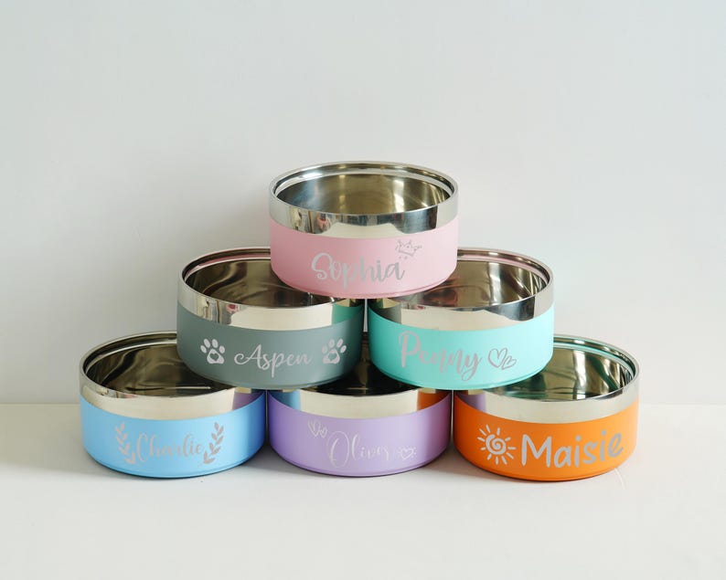 May include: A stack of six stainless steel pet bowls with colored exteriors. Each bowl has a different color, including pink, teal, blue, purple, and orange. The bowls are personalized with names and decorative icons.