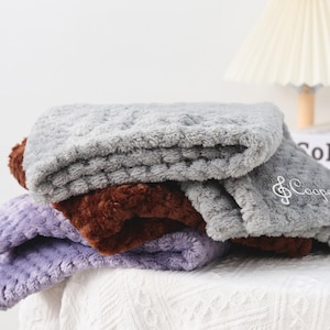 May include: A pile of three soft blankets in grey, brown, and purple. The top grey blanket is embroidered with the name "Cooper" and a musical note. The blankets have a textured, fluffy appearance, suggesting a cosy feel for the home.