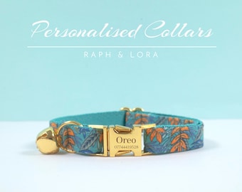 Puede incluir: Un collar para gato azul turquesa con una hebilla dorada y una campanilla. El collar tiene un patrón floral con hojas naranjas y azules. La hebilla tiene el nombre "Oreo" y un número de teléfono grabado.