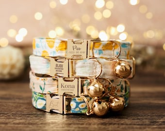 Puede incluir: Una pila de cuatro collares personalizados para gatos con estampados florales en amarillo, azul y rosa. Cada collar tiene una hebilla de metal dorado grabada con un nombre y un número de teléfono. Cuelgan campanas doradas de los collares.