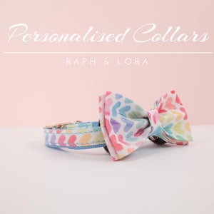 May include: A colorful pet collar with a rainbow heart pattern and a matching bow tie. The collar is blue with white stitching and the bow tie is made of the same fabric as the collar. The text "Raph & Lora" is printed on a pink background above the collar.