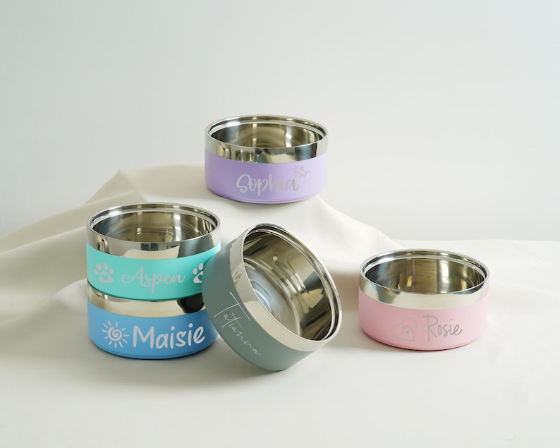 May include: Stainless steel pet bowls in various colors, including lavender, teal, and pink. Each bowl has a silver rim and is personalized with a pet's name in a script font. The bowls are stacked and arranged on a white surface.
