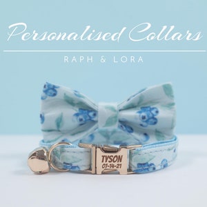 Puede incluir: Un collar de gato azul claro con una pajarita a juego con estampado de arándanos. El collar tiene una hebilla dorada con el nombre "TYSON" y la fecha "07-14-21" grabada.