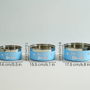 May include: Three stainless steel pet bowls with light blue bands. Each bowl has the name "NICOLE", a bone graphic, and paw prints. The bowls measure 5.5, 6.1, and 6.8 inches in diameter.