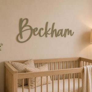 May include: A wall-mounted wooden sign featuring the name Beckham in a cursive script font. The lettering is finished in a muted sage green colour and is positioned horizontally above a light-toned wooden crib with a white mattress and pillows.