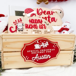 Puede incluir: Una caja de madera con un letrero rojo y dorado que dice "Entrega especial de Navidad para Austin". En el interior hay decoraciones navideñas, incluyendo un gorro de Papá Noel, un muñeco de nieve y una lista de deseos.