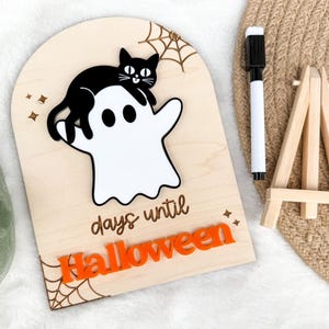 K&ouml;nnte beinhalten: H&ouml;lzernes Halloween-Countdown-Schild mit einem wei&szlig;en Geist und einer schwarzen Katze. Das Schild enth&auml;lt den Text "days until Halloween" und orangefarbene Akzente. Ein schwarzer Marker und eine kleine Holzstaffelei sind ebenfalls vorhanden.