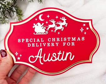 以下が含まれることがあります： 金色の縁取りが付いた赤いクリスマスサイン。サンタのそりとトナカイの白いイラストが描かれています。サインには白い文字で「SPECIAL CHRISTMAS DELIVERY FOR Austin」と書かれています。お祝いの休日の装飾。