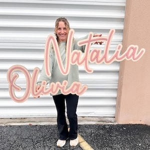 May include: Wooden sign featuring the names "Natalia" and "Olivia" in a layered design. The names are in a cursive font with a light pink outline and a natural wood interior. The sign is held up in front of a white corrugated metal background.