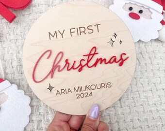 May include: A round wooden sign with the text "My First Christmas" in red cursive lettering. The sign also includes the name "Aria Milikurous" and the year "2024".