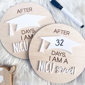 May include: Two wooden round signs with a white graduation cap on top. The signs are decorated with the text "After" and "Days, I am a NICU grad". The sign on the left says "After Days, I am NICU".