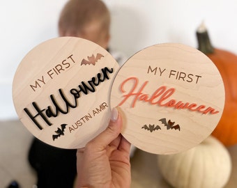 Peut inclure: Deux pancartes rondes en bois avec le texte "My First Halloween" en lettres brunes et orange. Une pancarte porte le nom "Austin Amir" écrit sous le texte. Les deux pancartes ont des découpes de chauves-souris noires.