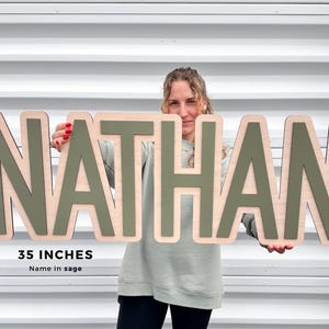 Op de afbeelding: Houten bord met de naam "NATHAN" in saliegroene letters, 89 cm breed. De letters zijn gemonteerd op een natuurlijke houten basis. Het bord wordt door een persoon vastgehouden.