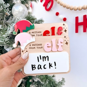 Peut inclure: Une pancarte d'elfe en bois avec un chapeau d'elfe rose et rouge, un tableau blanc et le texte "Un message de votre elfe" et "Je suis de retour !"