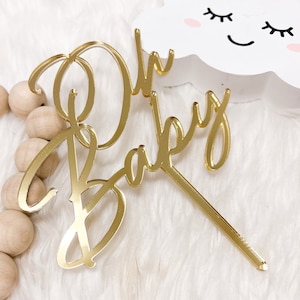 May include: Gold mirror acrylic cake topper with the words "Oh Baby" in a cursive script font. The topper is on a white fuzzy surface.