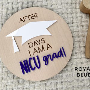 May include: A wooden round sign with the text "After days, I am a NICU grad!" in blue lettering. A white graduation cap is on top of the text. The sign is on a white fuzzy surface.