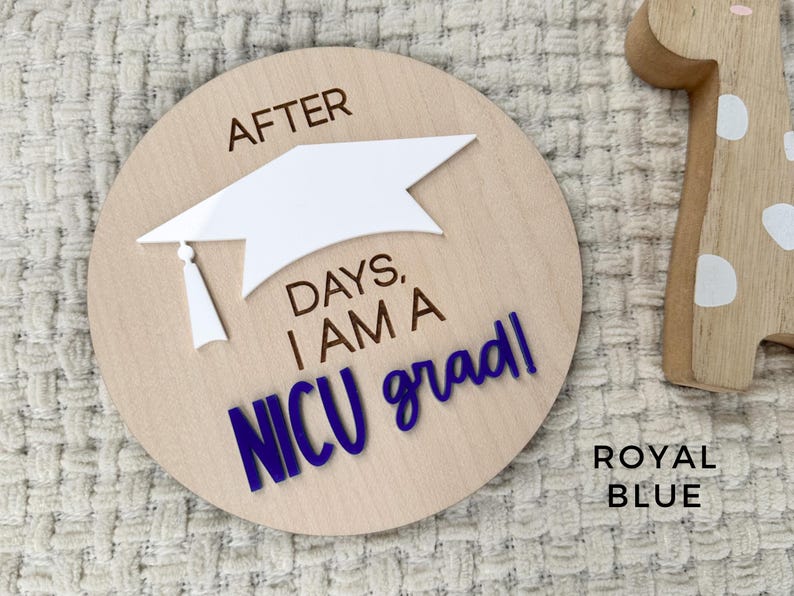 May include: A wooden round sign with the text "After days, I am a NICU grad!" in blue lettering. A white graduation cap is on top of the text. The sign is on a white fuzzy surface.