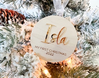 Puede incluir: Un adorno redondo de madera con el nombre "Isla" escrito en letras doradas. El adorno también tiene el texto "My First Christmas 2022" escrito en él.