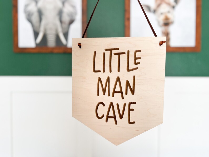 May include: A wooden banner with the words "LITTLE MAN CAVE" engraved in brown. The banner is light-colored and hangs from a brown leather cord. The background includes framed animal prints.