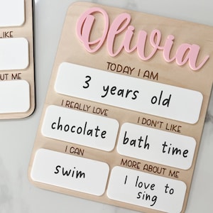 May include: A light-colored wooden board featuring the name Olivia in raised pink script. The board includes white rectangular sections with black text reading: Today I am 3 years old, I really love chocolate, I don't like bath time, I can swim, and more about me I love to sing.
