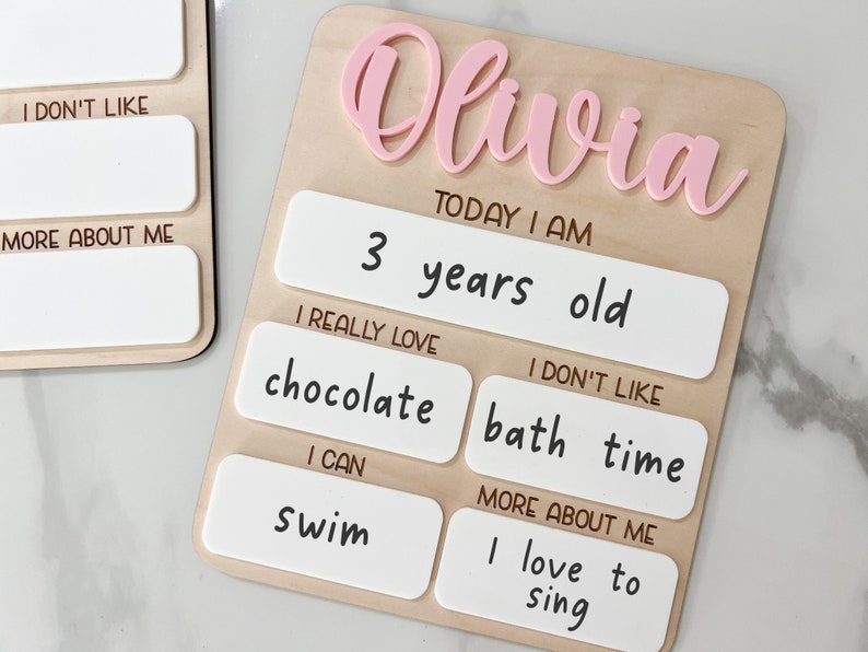 May include: A light-colored wooden board featuring the name Olivia in raised pink script. The board includes white rectangular sections with black text reading: Today I am 3 years old, I really love chocolate, I don't like bath time, I can swim, and more about me I love to sing.
