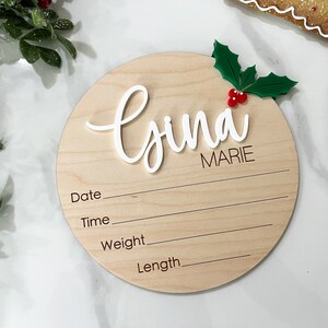 May include: A round wooden plaque with a white cursive name "Gina Marie" and a holly sprig with red berries. The plaque has lines for recording the date, time, weight, and length.